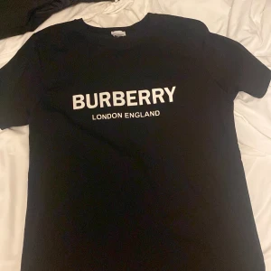 Svart Burberry t-shirt med tryck - Säljer en svart t-shirt från Burberry med klassiskt vitt tryck på bröstet där det står 'BURBERRY LONDON ENGLAND'. T-shirten har rund hals och korta ärmar. Perfekt för dig som gillar stilrena och ikoniska plagg.