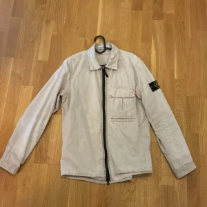 Beige overshirt från Stone Island - Snygg beige overshirt från Stone Island med klassisk krage, bröstficka och svart dragkedja framtill. Jackan har lång ärm och ikonisk logotyp-patch på vänster arm. Perfekt för lager-på-lager och stilren vibe Pris kan diskuteras vid snabb affär