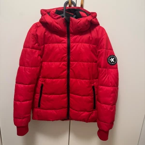 Röd pufferjacka från Everest - Riktigt snygg röd pufferjacka från Everest med huva och svarta detaljer. Jackan har dragkedja framtill, två fickor med dragkedja och en patch på ärmen. Perfekt för kyliga dagar och enkel att matcha med din stil.