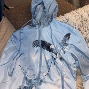 Blå vindjacka från Trapstar med örntryck - Snygg ljusblå vindjacka från Trapstar med stora örntryck både fram och bak samt broderad Trapstar-logga på ryggen. Jackan har huva med snörning, dragkedja framtill och två fickor med dragkedja. Perfekt statement-plagg för dig som vill sticka ut.