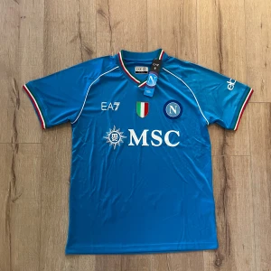 Napoli EA7 blå fotbollströja M - Napoli fotbollströja från EA7 i blått med vita detaljer och klubbmärke på bröstet. V-ringad krage med italienska flaggans färger, korta ärmar och tryckta sponsorer. Tillverkad i lätt polyester som andas, perfekt för match eller träning.