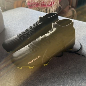 Nike Mercurial Air Zoom fotbollsskor - Säljer ett par svarta Nike Mercurial Air Zoom fotbollsskor med högt skaft och snörning. Skorna har en strömlinjeformad design med subtila silverdetaljer och gula dobbar. Materialet är syntet och skon är lätt och smidig för snabba rörelser på planen. Säljer dessa eftersom de inte passade mig.