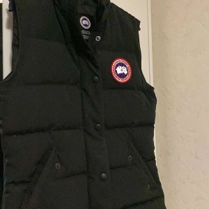 Svart dunväst från Canada Goose XS - Svart dunväst från Canada Goose i storlek XS. Västen har ett quiltat mönster, tryckknappar och dragkedja framtill samt den klassiska röda och vita logotypen på bröstet. Fick den i present men då de inte är min stil så har jag aldrig använt den, kvitto finns på denna, pris går att diskutera 