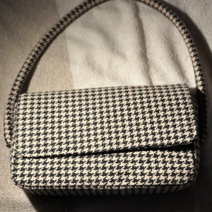 Rutig axelväska i beige och svart - Snygg axelväska med klassiskt hundtandsmönster i beige och svart. Väskan har en rektangulär form, bred axelrem och magnetlås. Materialet är mjukt och känns som bomull, perfekt för att lyfta din outfit med lite retrovibe.