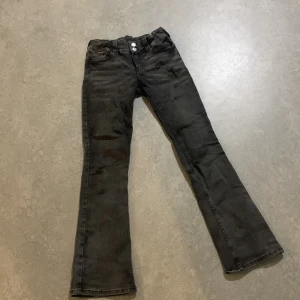 Svarta bootcut jeans med ficklock - Snygga svarta jeans med bootcut passform och coola ficklock med silverknappar bak. Jeansen har klassisk femficksdesign, två knappar i midjan och lätt tvättad look. Perfekta för dig som gillar en lite retro vibe och vill sticka ut med detaljer.