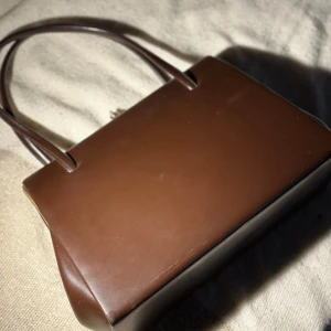 Brun vintage handväska  - Snygg brun handväska med stilren, rektangulär form och två smala handtag. Väskan är gjord i slätt skinn och har ett elegant guldfärgat lås upptill. Insidan är fodrad i matchande brun textil. Perfekt för dig som gillar retrovibbar och minimalistisk design.