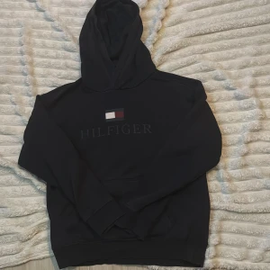Svart hoodie från Tommy Hilfiger - Svart hoodie från Tommy Hilfiger med klassisk logga och broderad text på bröstet. Tröjan har huva, lång ärm och en stor magficka framtill. Perfekt för dig som gillar stilrena och bekväma plagg med streetkänsla.