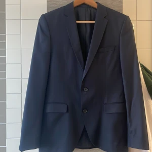 Mörkblå kavaj med två knappar - Snygg mörkblå kavaj med klassisk passform och två knappar framtill. Kavajen har smala slag, två fickor med lock och en bröstficka. Perfekt för dig som vill ha en stilren och tidlös look.