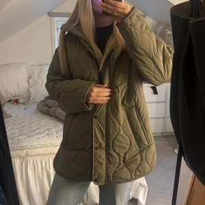 Olivgrön quiltad pufferjacka - Säljer en olivgrön pufferjacka med quiltad design och dragkedja framtill. Jackan har en oversized passform, hög krage och tryckknappar. Perfekt för kyliga dagar och riktigt snygg till jeans eller cargo. Materialet är syntet och jackan har långa ärmar.