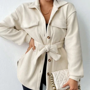 Beige overshirt med bälte och knappar - Stilren beige overshirt med knappar framtill, två bröstfickor och ett brett bälte i midjan som knyts. Jackan har lång ärm.                                              Kostade 330     