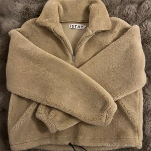Beige teddytröja från Stay, storlek S - Mysig beige teddytröja från Stay med halv dragkedja och hög krage. Tröjan är tillverkad i mjuk fleece och har en relaxed passform.