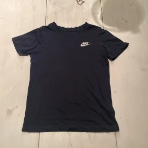 Mörkblå Nike t-shirt i bomull - Snygg mörkblå t-shirt från Nike med klassisk vit logga på bröstet. T-shirten är i mjuk bomull och har en enkel, stilren design med rund hals och korta ärmar. Perfekt för en sportig och avslappnad look.