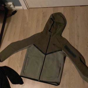 Olivgrön Nike hoodiejacka med dragsedia - Nike hoodiejacka i olivgrönt med svarta detaljer och hel dragkedja framtill. Jackan har huva, två sidofickor och en extra ficka med dragkedja på ärmen. Tillverkad i mjukt syntetmaterial, perfekt för en sportig och avslappnad stil.