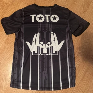 Svart Jul x Marseille fotbollströja - Snygg svart fotbollströja med gråa och vita vertikala ränder, tryck från Jul och Marseille, samt Yamaha-logga. Tillverkad i lätt och ventilerande polyester, perfekt för match eller träning. Passar även S i storleken.