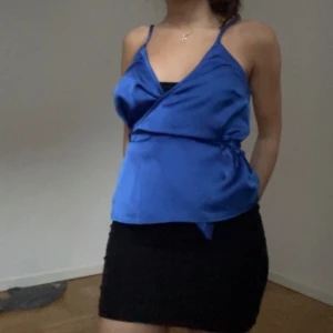 Blå omlott linnetopp i satin - Säljer en snygg blå omlott linnetopp med smala axelband och v-ringning. Toppen har en glansig satinfinish och knyt i sidan för en cool detalj. Perfekt för dig som gillar en stilren och trendig look med lite extra shine. 