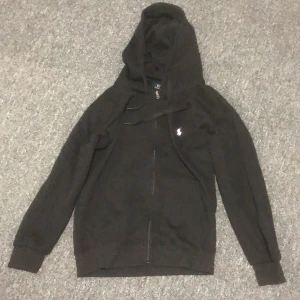 Svart hoodie från Polo Ralph Lauren - Svart hoodie från Polo Ralph Lauren i storlek S. Tröjan har huva, dragkedja framtill och långärmade ribbade muddar. Klassisk vit broderad logga på bröstet. Perfekt för chill dagar och enkel att matcha med jeans eller joggers.