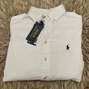 Vit slim fit skjorta från Polo Ralph Lauren - Vit skjorta från Polo Ralph Lauren i slim fit-modell med klassisk krage och knappar framtill. Broderad marinblå logga på bröstet. Tillverkad i mjuk bomull, perfekt för en clean och stilren look. Långärmad och med snygga detaljer som button-down krage. Har en pytte liten röd fläck på armen men märks absolut inte från långt håll. Använd en gång bara med lappar fof på 