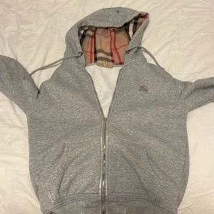 Grå hoodie från Burberry  - Grå hoodie från Burberry med klassiskt rutigt foder i huvan och broderad logga på bröstet. Tröjan har hel dragkedja, kängurufickor och justerbar huva. Tillverkad i mjuk bomullsmix, perfekt för chill dagar. Notera en defekt på dragkedjan som går att laga 