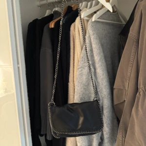 Svart axelväska från Gina Tricot - Snygg svart axelväska från Gina Tricot i skinnimitation med kedjedetaljer längs kanterna och som axelrem. Väskan har en mjuk, slät yta och minimalistisk design som ger en cool vibe. Perfekt storlek för det viktigaste och passar till många olika outfits.