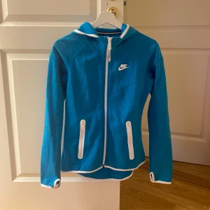 Blå Nike hoodie - Nike hoodie med vita detaljer, fickor med dragkedja, huva och hål till tummarna som värmer händerna. Storlek S