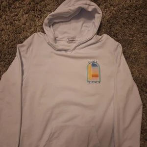 Vit hoodie från Casablanca med tryck - Vit hoodie från Casablanca med färgglatt tryck av ett fönster och texten 'Casa Blanca' på bröstet och ryggen. Tröjan har huva och känguruficka, perfekt för dig som gillar stilrena men ändå unika plagg.