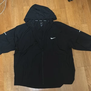 Nike Jacka - Svart vindjacka från Nike med huva och hel dragkedja framtill. Jackan har reflekterande detaljer på bröstet och ärmarna samt Nike-logga på bröstet. Tillverkad i lätt polyester, perfekt för löpning eller utomhusaktiviteter. Snygg och sportig look. StorlekX men jätte liten i storlek så perfekt för nån med  L.
