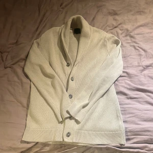 Beige kofta från H&M med knappar - Snygg beige kofta från H&M i storlek S, tillverkad i 100% bomull. Koftan har långärmad design, bred krage och knäppning med fyra knappar framtill. Ribbstickade muddar vid ärmslut och nederkant ger en clean och stilren look.