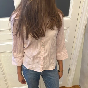 Ljusrosa skjorta med knappar - Snygg ljusrosa skjorta med krage och knappar framtill. Skjortan har ett subtilt randigt mönster och trekvartsärmar med breda manschetter. Materialet känns lätt och bomulligt, perfekt för dig som gillar en chill och fräsch stil.