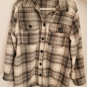 Rutig overshirt i grått och beige - Overshirt i mjukt material med rutigt mönster i grått, svart och beige. Skjortan har klassisk krage, knäppning framtill med svarta knappar och två bröstfickor med lock. Perfekt för lager-på-lager och chill stil.