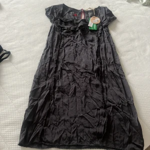 Svart Emelie Dress från Odd Molly - Snygg svart Emelie Dress från Odd Molly i färgen asphalt. Klänningen har korta ärmar, vackra spetsdetaljer och en feminin siluett. Tillverkad i ett glansigt material som ger en lyxig känsla. Perfekt för dig som vill ha en stilren och cool look. Klänningen är aldrig använd och därför skrynklig. Vid köp så stryks klänningen