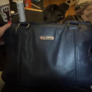 Mörkblå axelväska från Puccini - Snygg mörkblå axelväska från Puccini i skinnimitation med flera dragkedjefack och långa axelband. Väskan har stilrena metalldetaljer och en klassisk, rymlig design som passar perfekt för dig som vill ha både stil och funktion.