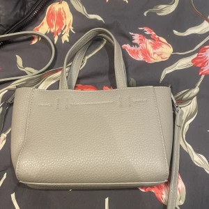 Ljusgrå axelväska i skinn - Snygg ljusgrå axelväska i strukturerat skinn med både handtag och avtagbar axelrem med kedjedetalj. Väskan har stilren design och dragkedja upptill. Perfekt storlek för det viktigaste och passar till många outfits.