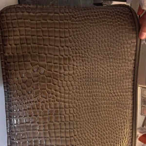 Brun datorväska i krokopräglat skinn - Snygg datorväska i brun färg med croco-präglat mönster och mjukt foder på insidan. Väskan har dragkedja runtom och passar perfekt för att skydda din laptop med stil. Materialet är syntetiskt skinn med en lyxig känsla.