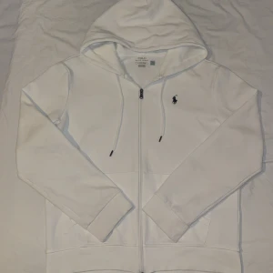 Vit hoodie från Polo Ralph Lauren - Vit hoodie från Polo Ralph Lauren med dragkedja och huva. Klassisk design med snörning i huvan och broderad logga på bröstet. Tillverkad i mjuk bomull, perfekt för en clean och stilren look.