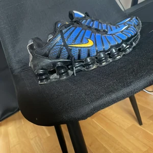 Nike Shox sneakers blå och svart - Nike Shox sneakers i blått och svart med gula swoosh-detaljer. Skorna har en unik fjädrande sula med Shox-teknologi och snörning. Ovandelen är i mesh och syntetmaterial med svarta plastdetaljer som ger en sportig vibe. Perfekt för dig som gillar retro och tech-inspirerad stil.