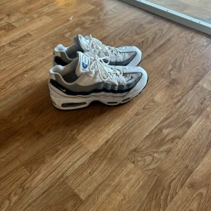 Nike Air Max 95 vita och blå sneakers - Nike Air Max 95 sneakers i vitt och marinblått med klassisk vågig design och synliga Air-enheter i sulan. Skorna har snörning, rund tå och platt sula. Materialmix av syntet och mesh för skön komfort och snygg streetstil. Perfekt för dig som gillar retrovibbar och sportig look. pris ej hugget i sten