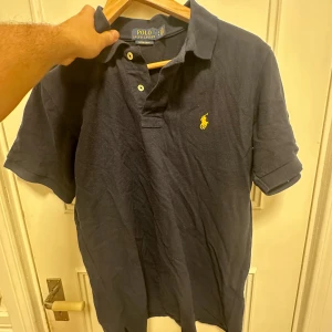 Mörkblå Polo Ralph Lauren piké - Snygg mörkblå pikétröja från Polo Ralph Lauren i slim fit-modell. Klassisk krage, korta ärmar och två vita knappar framtill. Ikonisk gul broderad logga på bröstet. Tillverkad i mjuk bomull som är skön mot huden.
