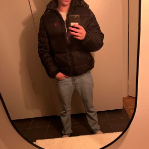 Svart pufferjacka Polo Ralph Lauren M - Svart pufferjacka från Polo Ralph Lauren i storlek XL men sitter som medium. Jackan har huva, dragkedja framtill, bröstficka med dragkedja och klassisk röd Polo-logga på bröstet. Tillverkad i syntetmaterial och perfekt för kalla dagar. Priset går att diskutera vid snabb affär 