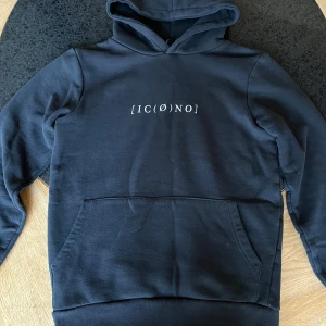 Svart hoodie från Icono XS - Svart hoodie från Icono i regular fit med vit textprint både fram och bak. Tröjan har huva, magficka och ribbade muddar. Materialet är mjuk bomull och polyester, perfekt för chill dagar. Snygg och enkel design med coolt tryck.