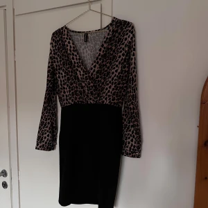 Leopardmönstrad klänning från New Collection - Snygg kort klänning med leopardmönstrad överdel och svarta kjoldel. V-ringad omlott upptill, långa ärmar med knappdetalj vid mudden. Använd max 2 gånger, så den är mycket fint skick! Storlek M