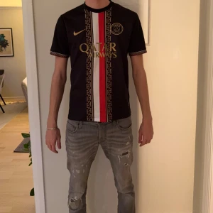 PSG fotbollströja Nike Dri-Fit L - Snygg PSG fotbollströja från Nike i storlek L. Tröjan är svart med guldiga detaljer, röd och vit rand i mitten samt Paris Saint-Germain-logga på bröstet. Tillverkad i lätt och ventilerande Dri-Fit material, perfekt för träning eller match.