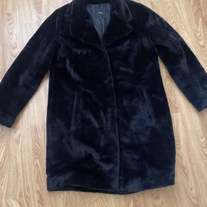 Svart pälsjacka från Object - Mysig svart pälsjacka från Object med klassisk krage och knäppning framtill. Jackan har två fickor på sidorna och är helfodrad för extra komfort. Perfekt för dig som vill ha en stilren och fluffig look under kyliga dagar.