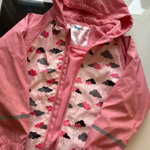 Rosa regnjacka med molnmönster Lupilu - Söt rosa regnjacka från Lupilu med huva och dragkedja. Jackan har ett gulligt mönster med moln och regndroppar i rosa och grått, samt reflexdetaljer på ärmarna. Insidan är fodrad med mjuk fleece för extra komfort.