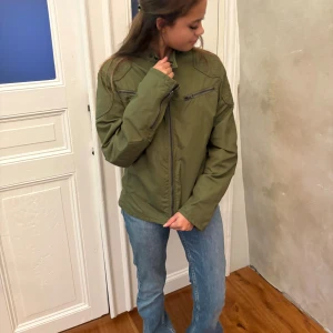 Grön vindjacka från Ralph Lauren - Säljer en grön vindjacka från Ralph Lauren med coolt emblem på ärmen och quiltade detaljer vid axlarna. Jackan har två snedställda bröstfickor med dragkedja, ståkrage och en snygg, lite loose passform. Perfekt för dig som gillar sportig och stilren look.