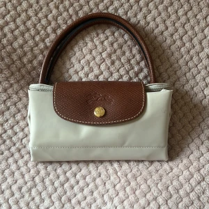 Beige handväska från Longchamp - Stilren beige handväska från Longchamp med bruna skinnhandtag och lock. Väskan har guldfärgad knapp och dragkedja, klassisk form och är tillverkad i syntetmaterial med detaljer i skinn. Perfekt storlek för att bära med sig det viktigaste.