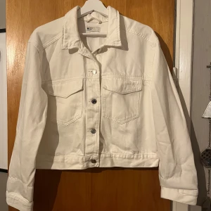 Vit jeansjacka från Gina Tricot - Vit jeansjacka från Gina Tricot med klassisk krage, två bröstfickor med lock och silverfärgade knappar framtill. Jackan har en rak, något croppad passform och långa ärmar. Strl L men funkar även som M