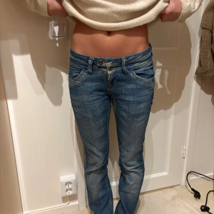 Blå lågmidjade jeans med brodyr - Snygga blå jeans med låg midja och bootcut-snitt. Jeansen har dubbla knappar i midjan och cool broderad detalj på bakfickorna. Klassisk femficksmodell i mjukt denimtyg, perfekt för en avslappnad stil.