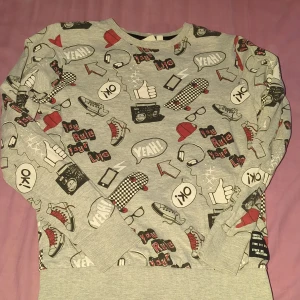 Grå sweatshirt med coolt tryck KappAhl - Grå sweatshirt från KappAhl med massor av grafiska tryck som sneakers, skateboards, kepsar och textbubblor i svart, rött och vitt. Tröjan har rund hals, långa ärmar och ribbade muddar. Perfekt för dig som gillar streetstyle och sköna vibes.