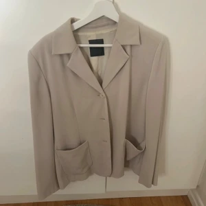 Beige kavaj från CCDK med fickor - Stilren beige kavaj från CCDK med klassisk krage, stora fickor framtill och knäppning med knappar. Jackan har en rak passform och är helfodrad, perfekt för en clean och snygg look.