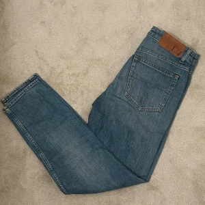 Blå jeansbyxor från tiger of Sweden 27/32 - Klassiska blå jeans med rak passform och femficksdesign. Byxorna har en brun TOS lapp bak i midjan och är tillverkade i slitstarkt denim. Perfekta för en avslappnad och tidlös look.